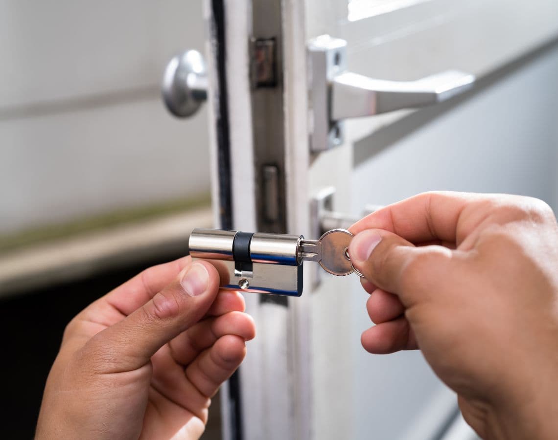 Manos instalando un cilindro de cerradura plateado con una llave en una puerta blanca.
